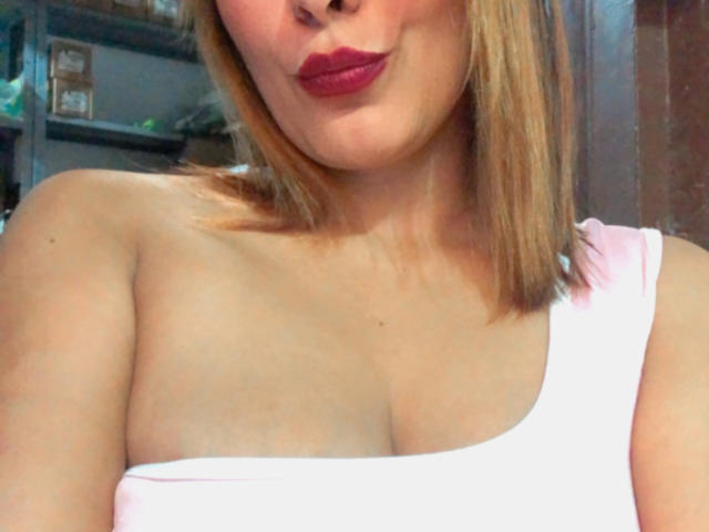 SaritaVelez met blond haar, volle, roze lippen, een wit topje met één schouderband dat haar borsten laat zien, lichte huid en subtiele make-up. Haar tepels zijn licht zichtbaar onder het topje..