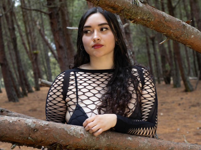 Antonnella Tyler, com cabelos longos e escuros, vestindo uma blusa de rede preta, expondo seus seios, está em uma floresta, segurando um galho de árvore, olhando para o lado e usando batom vermelho..