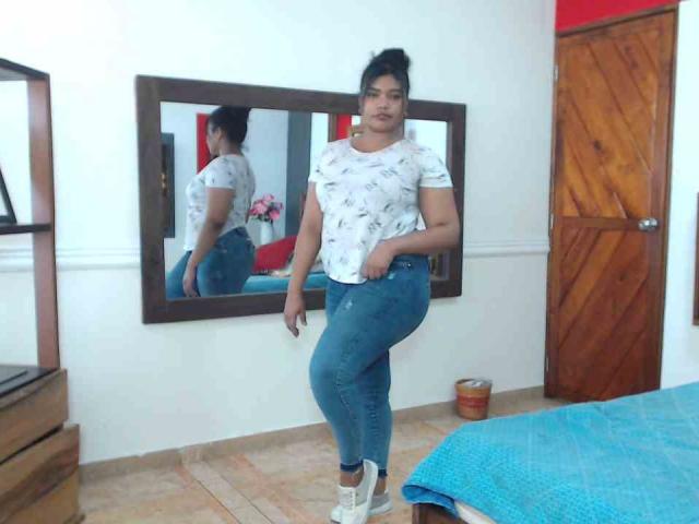 CataleyaX69 standing in a room, wearing a white patterned t-shirt, blue jeans, and white sneakers, with a mirror reflecting her ass. Her black hair is in a messy bun.