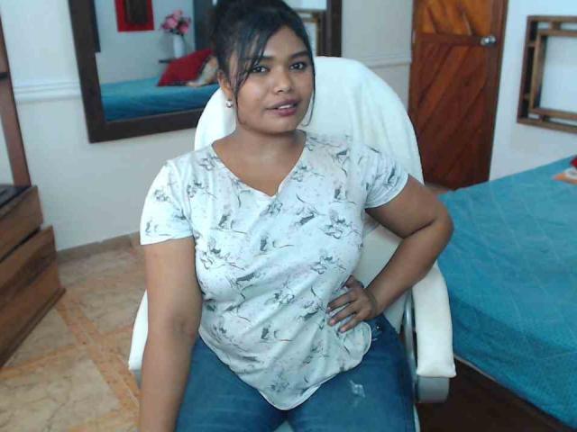 CataleyaX69 with dark hair in a ponytail, wearing a white floral shirt and blue jeans, sitting in a white chair, hand on hip, smiling, brown eyes, medium tits.