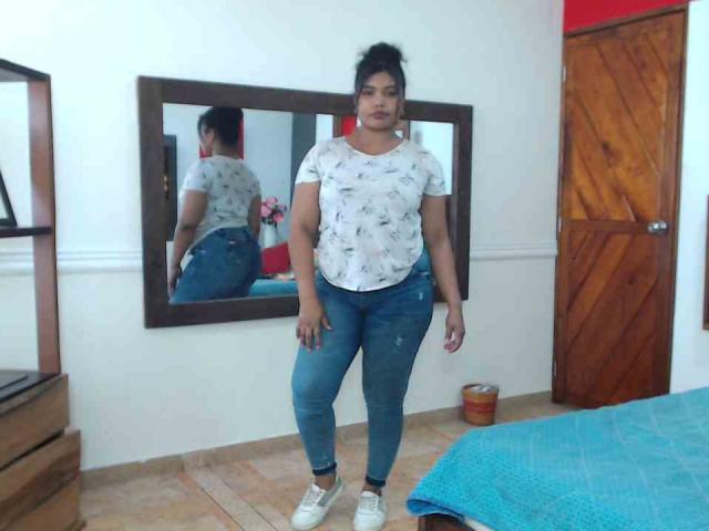 CataleyaX69 stands in a room, wearing a white floral shirt, blue jeans, and white sneakers. Her dark hair is in a messy bun, reflecting in a mirror.