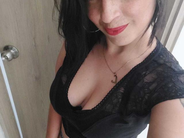 KarenBroc con una blusa de encaje negro, lápiz labial rojo, busto medio, y un collar con una cruz, sonriendo ligeramente, cabello negro y aros grandes, con un fondo de una puerta de madera..