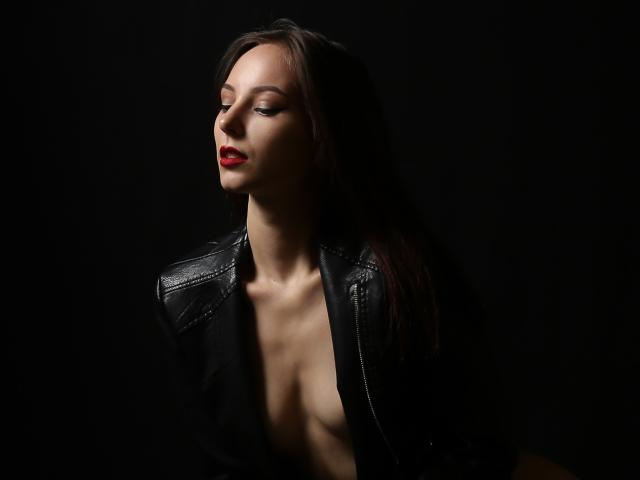 Aphelia Addington mit roten Lippen, dunkelbraunen Haaren, einer schwarzen Lederjacke und freiliegenden Brüsten, mit geschlossenen Augen, vor einem dunklen Hintergrund, wirkt sie sexy und selbstbewusst..