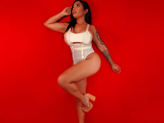 VickyGreys with large tits, wearing a white cut-out leotard, tan high heels, and tattoos, standing against a bright red background, her ass slightly raised, looking to the side.
