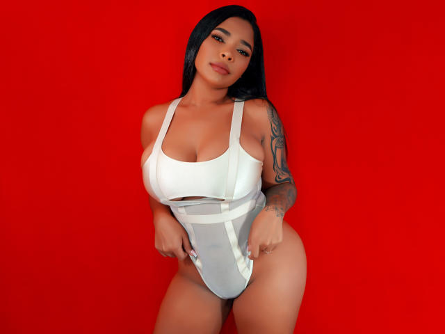 VickyGreys with black hair, large tits, and a tattoo on her right arm, wearing a white one-piece, pulling it down to show her pussy, against a red background.