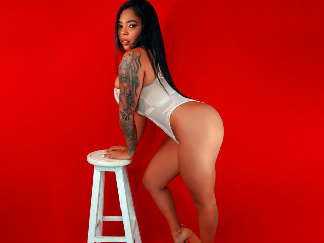 VickyGreys with long black hair, tattooed arm, wearing a white thong bodysuit, leaning on a white stool against a red background, showing her ass and looking over her shoulder.