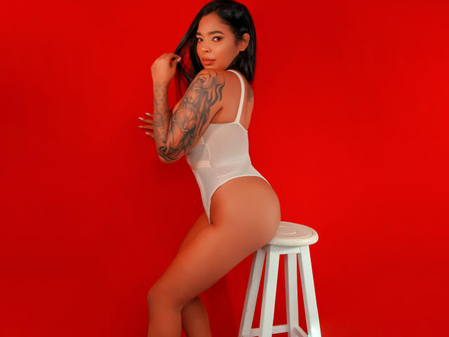 VickyGreys with tattoos, white sheer bodysuit, sitting on stool, red background, looking over shoulder, hand in hair, showing ass and tits, dark hair, medium skin, sexy.