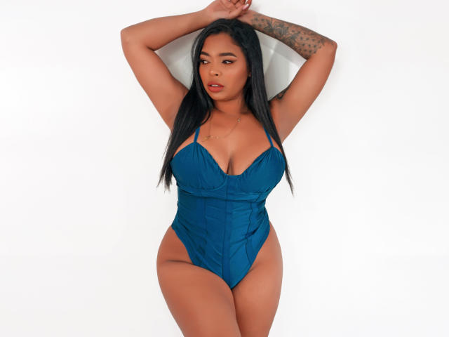 VickyGreys with long black hair, tan skin, and tattoos, wearing a blue, high-cut, one-piece, showing her big tits and curvy ass, arms raised, standing against a white wall.