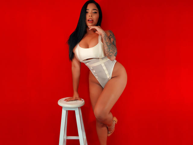 VickyGreys with long black hair, tattooed arm, and curvy body, wearing a white high-waisted thong, leaning on a white stool, red background, and tan high heels.