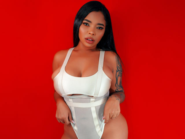VickyGreys with large tits, wearing a white one-piece, pulling it down slightly, showing a tattoo on her right arm, against a solid red background.