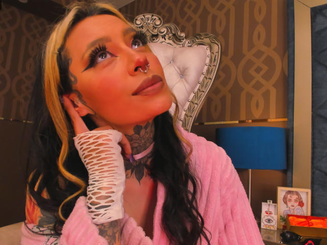 AlannaJade, con capelli biondi e neri, indossa un abito rosa, guanti bianchi senza dita e un anello al naso, guarda verso l'alto con il collo e la mano tatuati, e ha una mano sul viso..