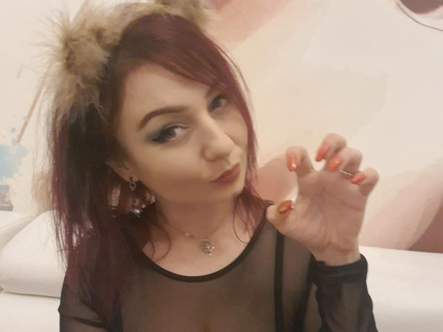JolieXKitty con i capelli rossi, orecchie da gatto e top in rete nera, con un'espressione accigliata e unghie arancioni, mostra l'anello che indossa, ha piccoli seni e porta una collana..