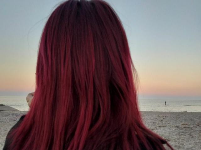 El cabello rojo, lacio y de longitud media de Zanoza cubre su espalda mientras mira una playa tranquila, iluminada por el atardecer, con una pequeña figura distante cerca de la orilla del agua..