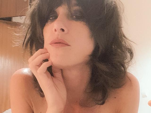 Aiyana with messy black hair, pale skin, and full lips, touching her chin with a hand, looking up with a slightly pouty expression, topless, in a soft-lit photo.