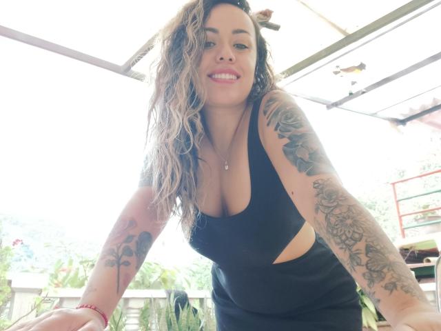 Joanallen, con capelli castano ondulati, canotta nera con aperture che mostra il seno, tatuaggi sulle braccia, sorridente, braccialetto rosso, in posizione leggermente inclinata in avanti, in un ambiente esterno luminoso..