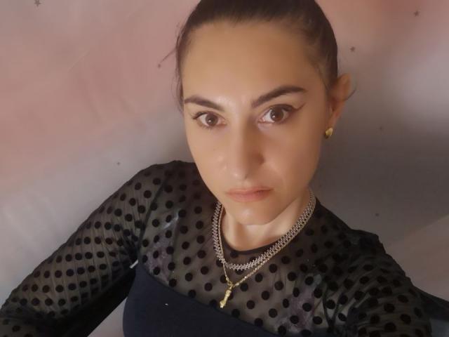 EvilFindom tiene ojos marrón oscuro, cejas arqueadas y lleva una camisa negra con lunares y un collar de cadena, mirando directamente a la cámara con una expresión seria..