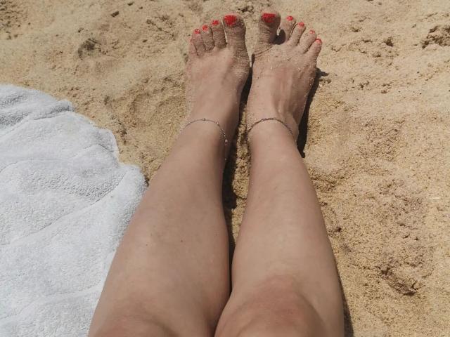 Los pies de EvilFindom, con los dedos de los pies pintados de rojo y adornados con tobilleras delgadas, están sobre una toalla de playa..
