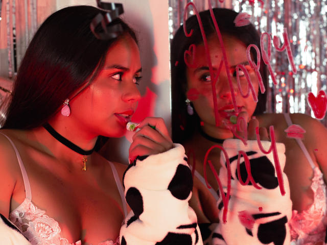 Arielle Wilde, avec ses longs cheveux noirs, portant un collier noir et un soutien-gorge en dentelle, mange des bonbons devant un miroir avec des lettres roses "AMOUR", et tenant une manche à motifs de vache..