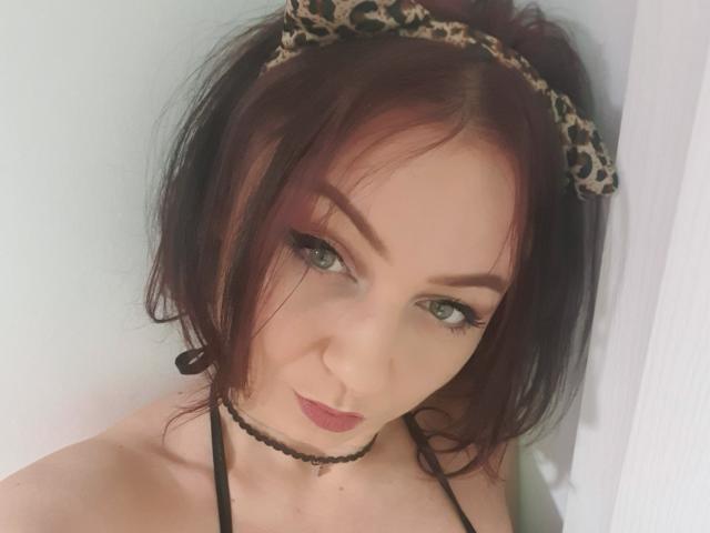 JolieXKitty with red hair, wearing a leopard headband, black choker, and eyeliner, looking up with green eyes, and a slight smirk, showing her bare shoulders.