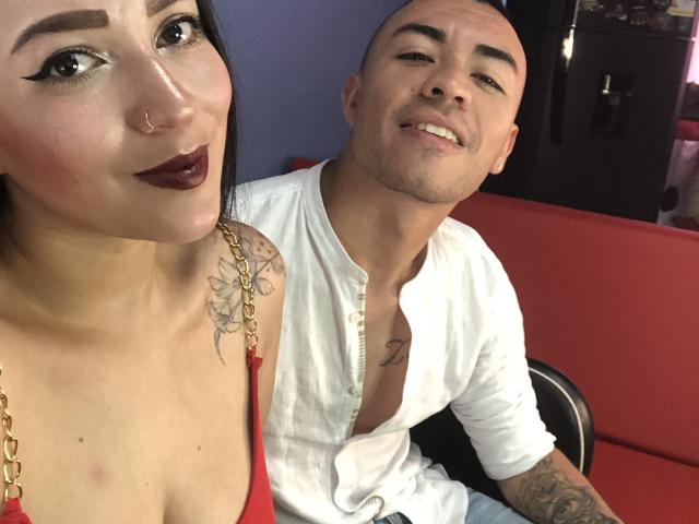 JaneAndPaull con rossetto scuro, piercing al naso e una maglietta rossa che mostra il seno, tatuaggio sulla spalla, seduta accanto a Paull, calvo, con una camicia bianca, entrambi sorridenti..
