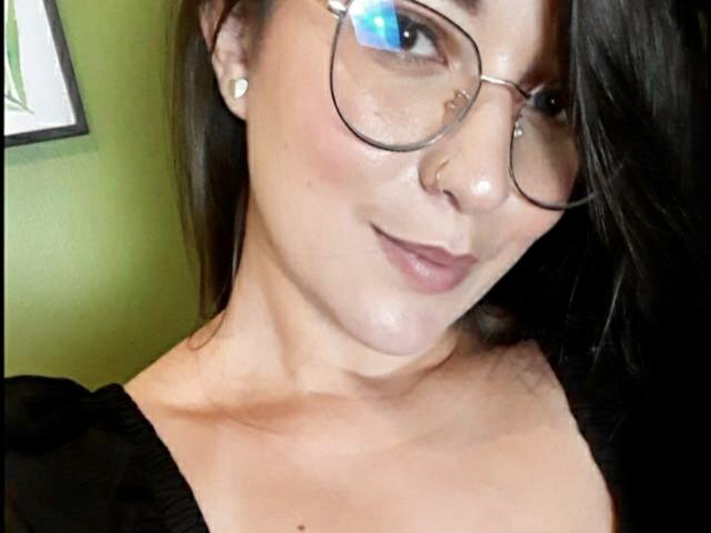 JaneAndPaull mit Brille, Nasenring und schwarzem Kleid, zeigt ein leichtes Lächeln, wobei ihre Brüste sichtbar sind. Sie hat helle Haut, braunes Haar und ein süßes Gesicht. In der Brille ist ein blauer Schimmer zu sehen..