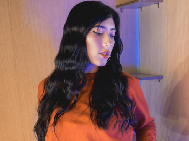 VioletaParr tiene el pelo largo, negro y ondulado, lleva una blusa naranja, con una iluminación azul tenue, un piercing en la nariz y delineador de ojos, está mirando hacia abajo con los ojos cerrados y tiene los labios ligeramente entreabiertos y de color rojo..