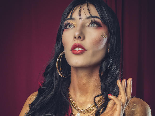 VioletaParr heeft zwart, golvend haar, rode lippenstift, gouden oorbellen en een gouden ketting. Ze kijkt omhoog met een lichte glimlach, haar hand in de buurt van haar gezicht. Rode achtergrond..