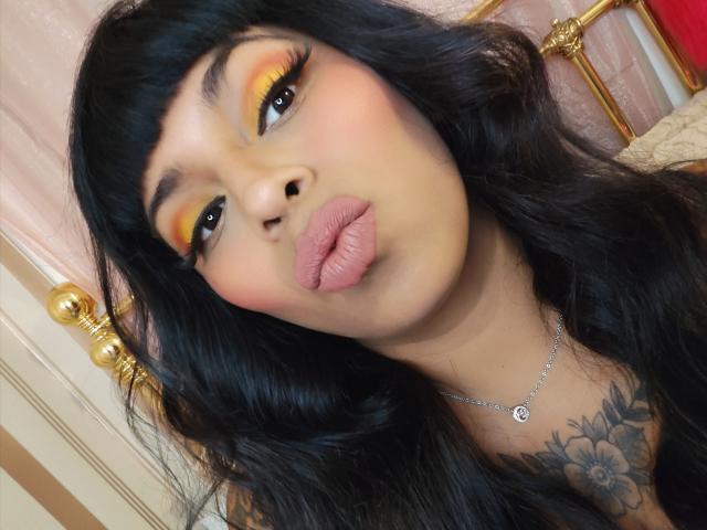 Chili is pouting with thick, pink, slightly glossy lips, her orange and yellow eyeshadow matching her seductive gaze, black tattoo on chest.