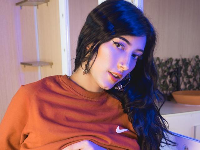 Violeta Parr, con lunghi capelli neri, indossa una maglietta arancione Nike, sollevandola per mostrare i suoi piccoli seni, guarda in basso con la pelle leggermente bluastra, orecchini a cerchio..