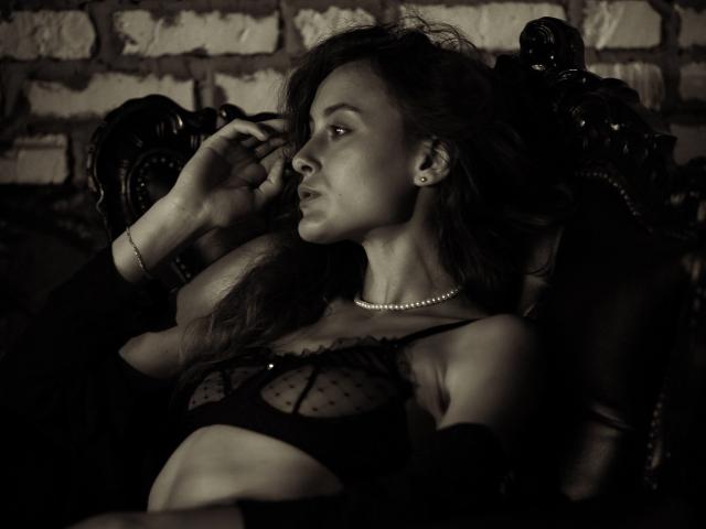 AlesyaDrons with dark hair, pearl necklace, and black mesh bra, sitting on an ornate chair, touching her ear, looking to the side, in a sepia-toned, sexy photo.