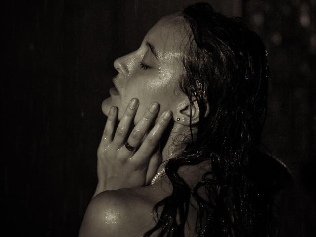 AlesyaDrons with wet, dark hair, eyes closed, hands on face, shiny skin, small stud earring, ring on finger, side profile, dark background, monochrome, sensual, water droplets.