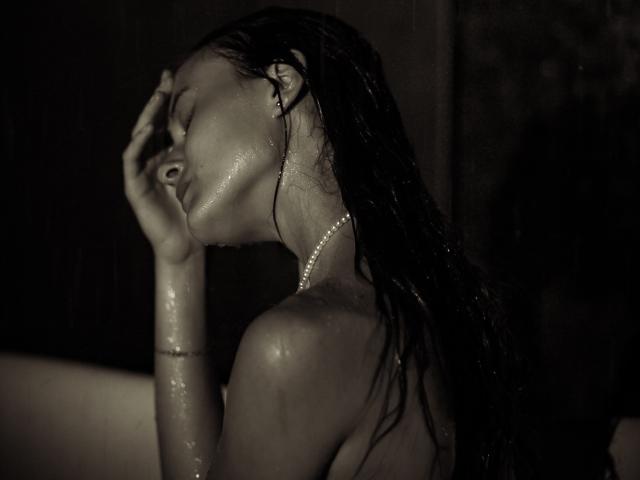 AlesyaDrons with wet, dark hair, eyes closed, hand on head, shiny skin, pearl necklace, and subtle bracelet, in a dark, rainy, sensual pose. Boobs visible, topless.