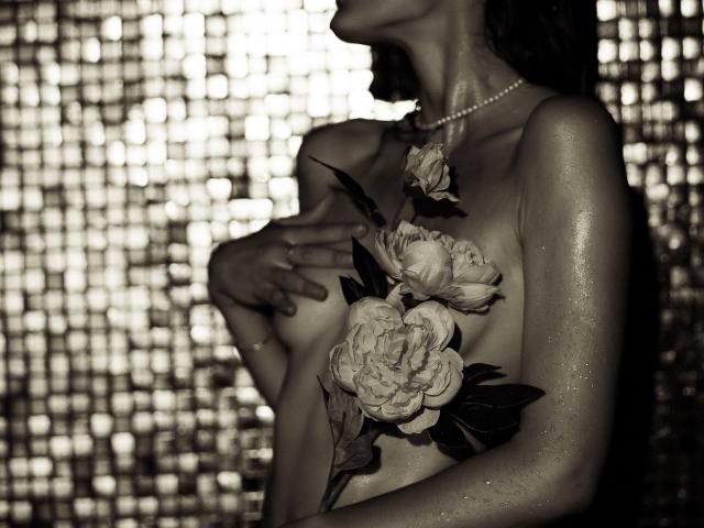 Alesya Drons, nue et avec une peau brillante, portant un collier de perles, a de grandes fleurs décoratives recouvrant ses seins, son bras levé, sa main sur sa poitrine, dans une photographie sombre, humide et sensuelle..