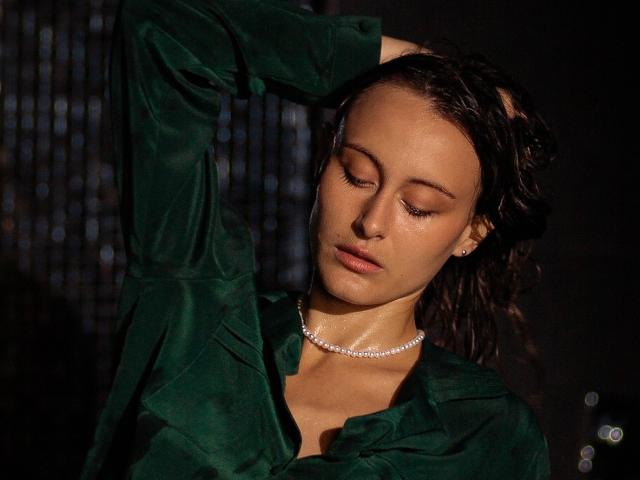 Alesya Drons, con gli occhi chiusi, indossa una maglietta verde e una collana di diamanti, e alza il braccio destro, mostrando capelli castani ondulati e pelle liscia e abbronzata. Il seno non è visibile..