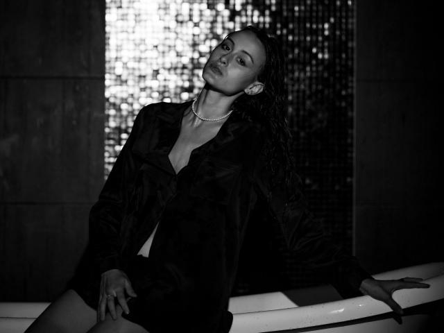 AlesyaDrons with wet, dark hair, wearing an open black shirt, revealing small tits, sitting in a white bathtub, looking up, in a dark, moody black-and-white photo.