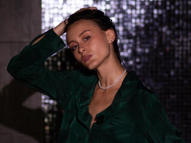 AlesyaDrons con i capelli castano scuri bagnati, una maglietta verde, una collana di perle, una mano tra i capelli, sexy, sicura di sé, un leggero sorriso, illuminazione soffusa, sfondo scuro e riflettente. Il seno è parzialmente visibile, la maglietta è aperta..