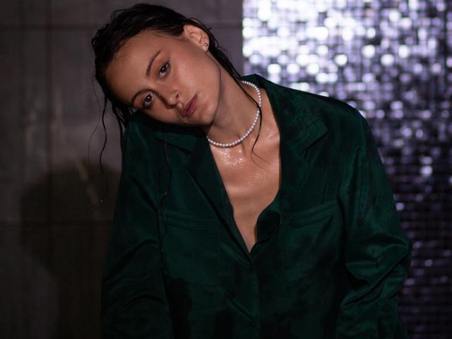 Alesya Drons, met nat, donker haar, die een groen, nat overhemd met knopen en een parelketting draagt, heeft een sexy, vermoeide uitdrukking en kijkt naar beneden met licht geopende lippen..