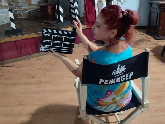 MilayaDFlow con i capelli rossi raccolti in una coda di cavallo, indossa una camicia blu e ha uno sfondo colorato, tiene in mano una clapperboard e siede su una sedia da regista con l'etichetta "PENNAGER". Pavimento in legno, orecchini viola..