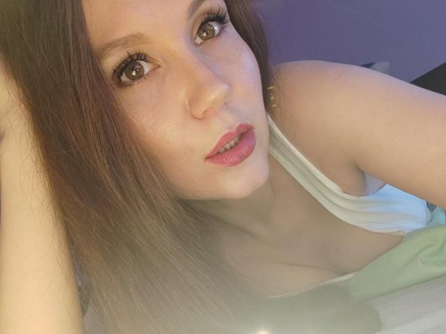 RittaSweetRose with long brown hair, pink lipstick, and brown eyes, wearing a white tank top, showing her small tits and pale skin, lying down with a relaxed expression.