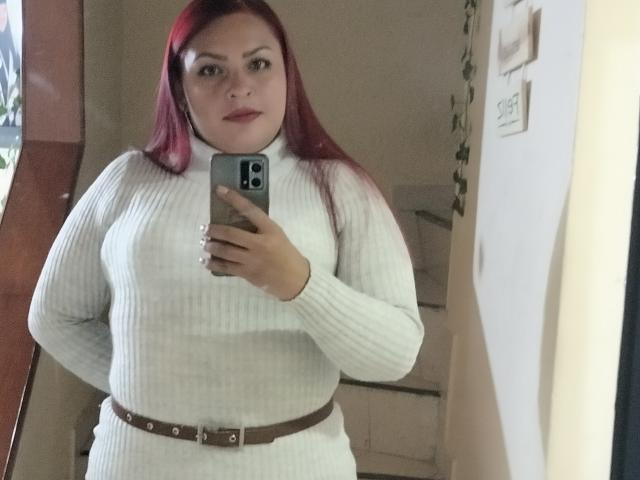 Adi69, avec des cheveux roux, portant un pull col roulé blanc texturé et une ceinture marron, prend un selfie devant un miroir, montrant ses seins et ses fesses. Elle tient un téléphone gris dans sa main droite..