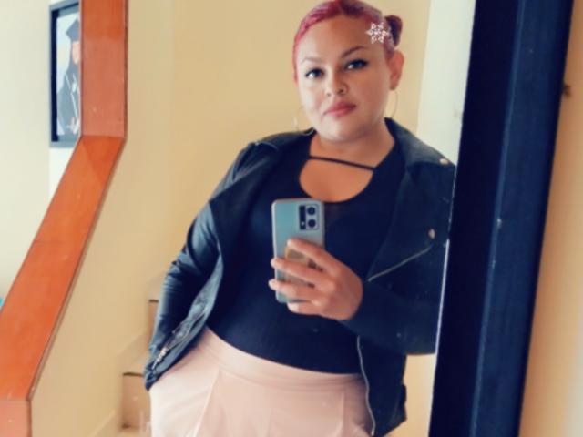 Adi69 con i capelli rossi raccolti in uno chignon, giacca di pelle nera, top nero, gonna rosa, che tiene in mano il telefono, selfie allo specchio, seno di media grandezza, labbra rosa, collana nera, riflesso nello specchio, sguardo sicuro..
