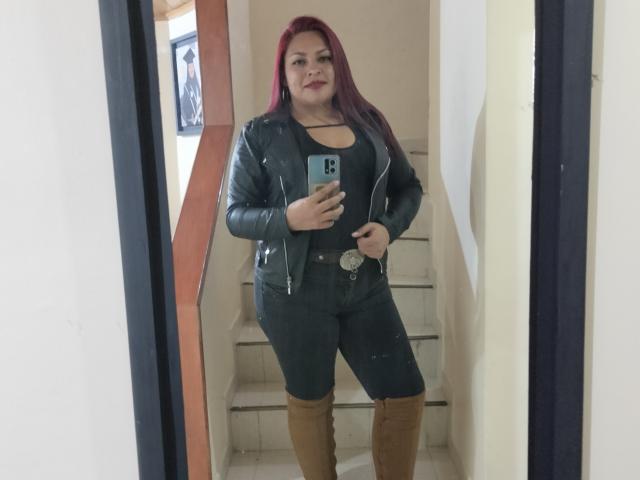 Adi69 con i capelli rossi, una giacca di pelle nera, una canottiera nera, pantaloni neri e stivali marroni, in piedi davanti a uno specchio, che tiene in mano un telefono, mostrando un sedere paffuto e formoso..