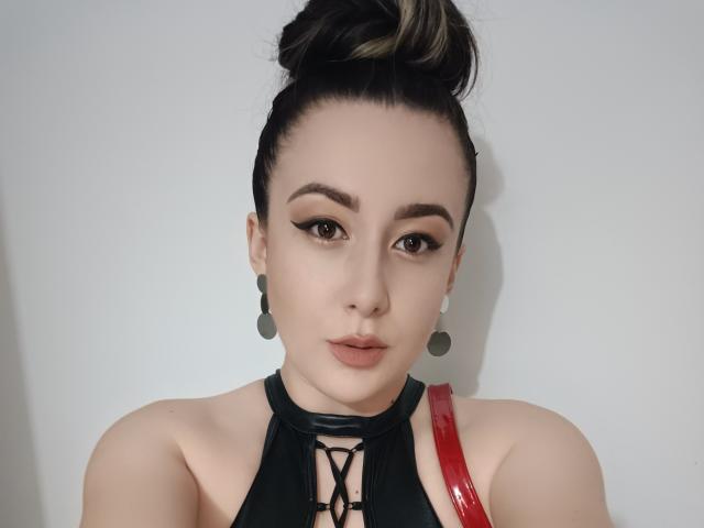 Maby Legrand mit dunklem Haar zu einem Dutt gebunden, trägt eine schwarze Spitzenbluse mit Schnürung, einen roten Träger, auffälligen Eyeliner und runde Ohrringe. Sie blickt mit neutralem Ausdruck in die Kamera..