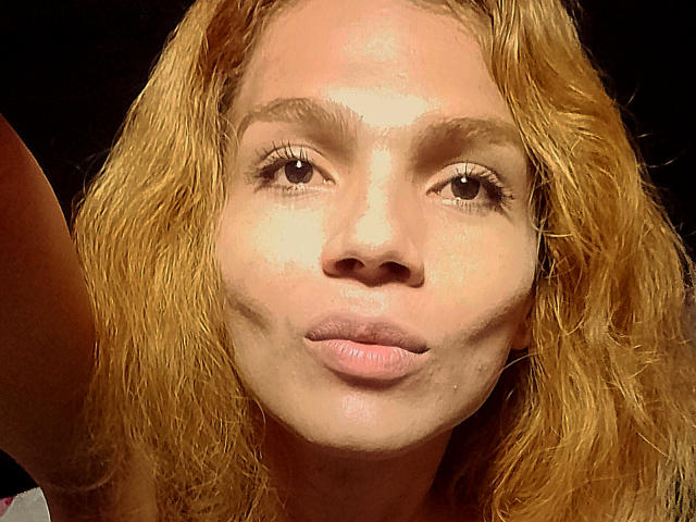 TatyLorenX hat welliges, blondes Haar, helle Haut, braune Augen und zieht die Lippen leicht zusammen, wobei sie einen rosafarbenen Lippenstift trägt. Ihr Gesicht ist zentriert, mit einem dunklen Hintergrund..