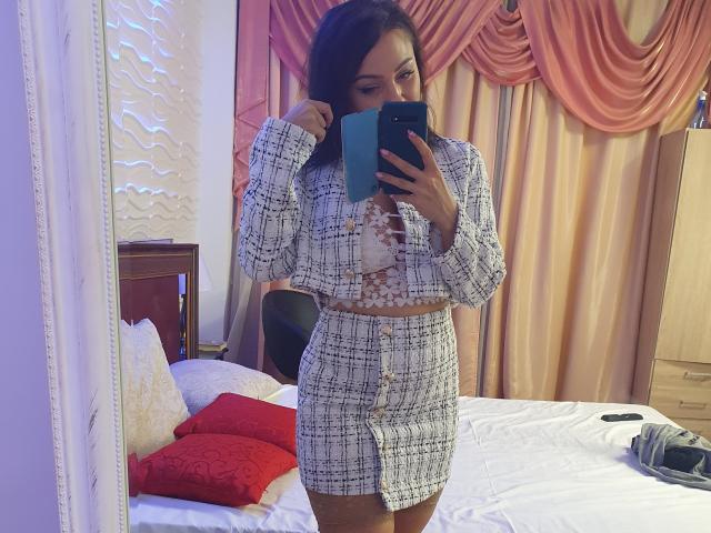 JolieXKitty in a plaid outfit with button-down front, showing tits in a white floral bra, taking a mirror selfie with a blue phone, in a bedroom.