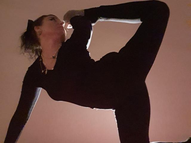 JolieXKitty is in a black outfit, doing a high kick with one leg raised, showing her flexible ass and strong legs, against a dimly lit background.