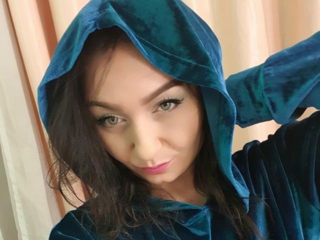 JolieXKitty with dark hair, green eyes, and pink lipstick, wearing a blue velvet hood, slightly smiling, hand in hood, soft light, looking at camera.