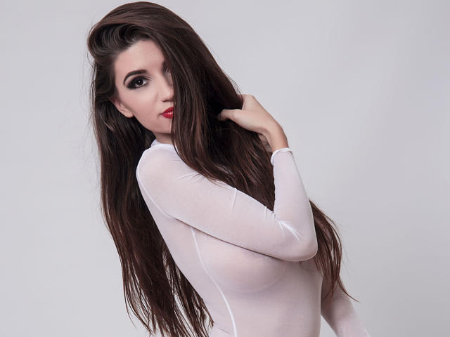 DeliciouseMila, com longos cabelos castanhos, lábios vermelhos, vestindo uma blusa branca transparente, mostrando os seios, com a mão no cabelo, olhando para a câmera, sexy, pele clara, fundo branco..
