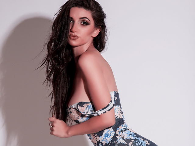 DeliciouseMila mit langen, dunklen Haaren, die ein trägerloses, geblümtes Kleid trägt, wobei ihre Brüste sichtbar sind, blickt über ihre Schulter mit leicht geöffnetem Mund und einem verführerischen Lächeln..