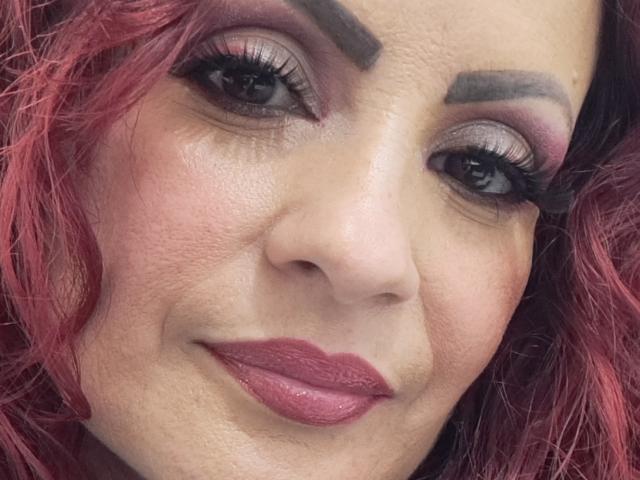 Das Gesicht von Karina Molina mit rosa-rotem Haar, dunklen Augenbrauen, starkem Eyeliner, langen Wimpern und rosa Lippenstift. Sie lächelt leicht und zeigt eine leicht faltige Hautstruktur..